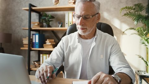 Middle-aged man in glasses and casual outfit is typing on notebook and writing down something while sitting at table working at home office. Remote job, freelance, business. Close up