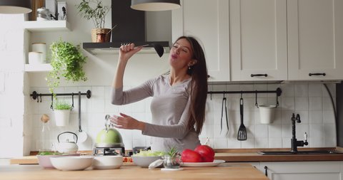 Happy relaxed young woman singing song in ladle as microphone, dancing alone in modern kitchen. Emotional active millennial girl having fun, distracted from preparing domestic morning healthy meal.