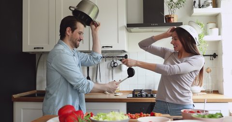 Overjoyed young family couple using kitchen ware utensils as helmets and swords playing fight battle in kitchen, happy spouses involved in childish activity, having fun spending weekend leisure time.