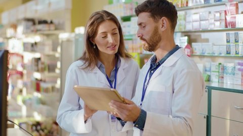 Male caucasian pharmacy intern exploring medications with digital tablet asking advice from professional apothecarist cooperating in drugstore. Healthcare and medicine.
