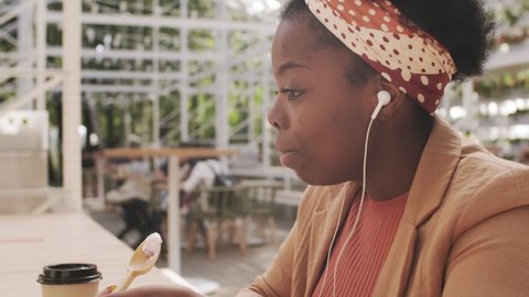 Side view shot of beautiful African-American woman in earphones sitting at table in outdoor patio of cafe and chatting on video call while enjoying ice cream on her lunch break