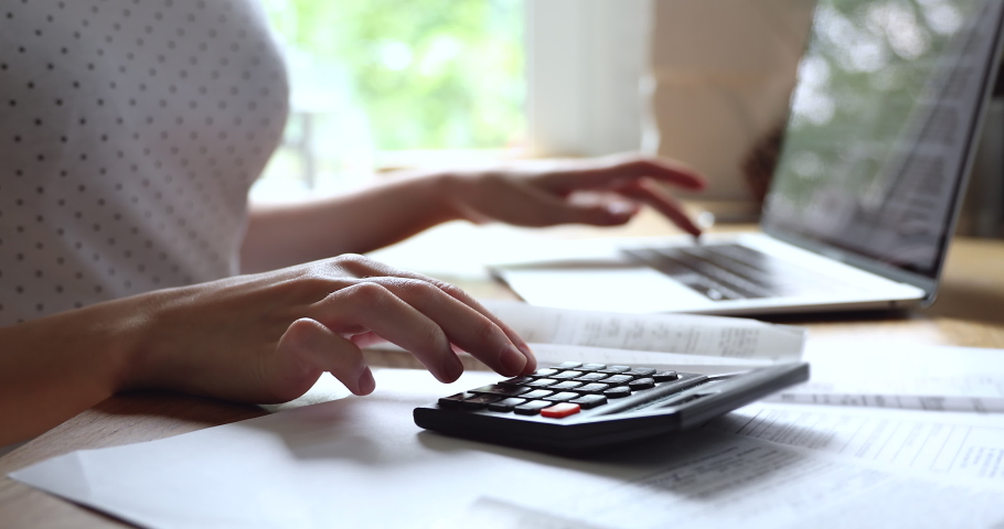 Close up young woman summarizing monthly incomes and outcomes with calculator, checking electronic bills in e-banking computer application, managing expenditures and household money payments.