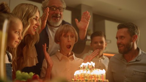 Big family celebrating grandmother birthday at home. Senior man closing woman eyes for surprise. Excited mature woman looking at cake with burning candles. Happy family clapping hands together