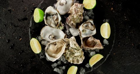 Oysters with pieces of lime and ice on stone board rotate. On black background.