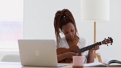 young african american woman is learning to play the ukulele guitar and checking chords  . using laptop computer. Girl is smiling and looking at screen, use the music instrument for hobby