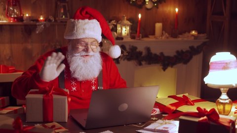 Santa Claus holding gift box video calling kid talking to child greeting on Merry Christmas, Happy New Year in virtual video online chat on laptop sitting at home table late with present on xmas eve.