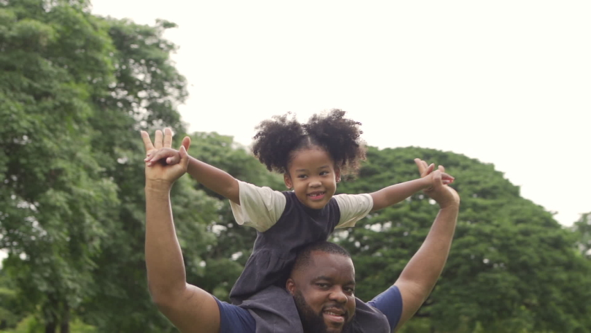 Happy affectionate mixed race family. African man father carrying little daughter on his back and walking in the park. Dad and cute child girl enjoy spending time together in outdoor weekend vacation. Royalty-Free Stock Footage #1061203660