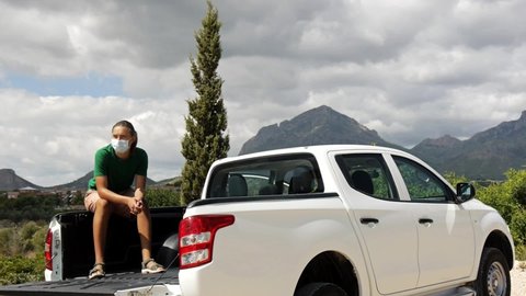 Teenager boy in protective mask sitting in back of pickup truck on mountain background in countryside.