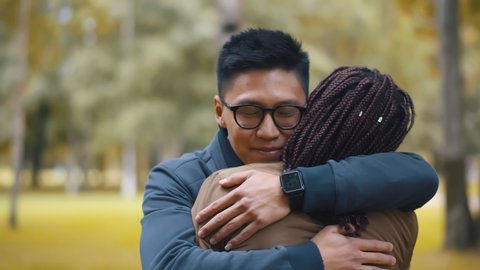 Diverse millennial couple meeting in autumn park and embracing each other. Back view of african woman greeting asian man with hug standing outdoors. Multiracial friendship and relationship concept