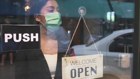 Business owner attractive young Asian woman in suit hanging we're open sign on front door  welcoming clients to new cafe. Happy waiter with protective face mask holding open sign  at hotel .