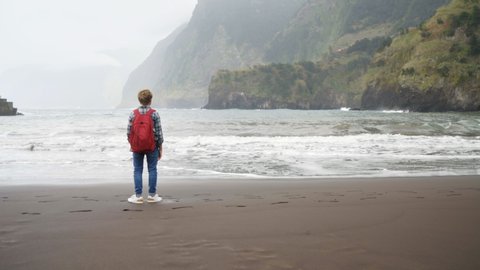 4K boy standing on black sandy beach watching ocean waves with backpack. Traveling concept.