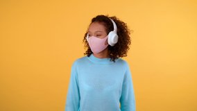 A woman in a protective medical mask and large white headphones is having fun and dancing. Isolated yellow background 4K