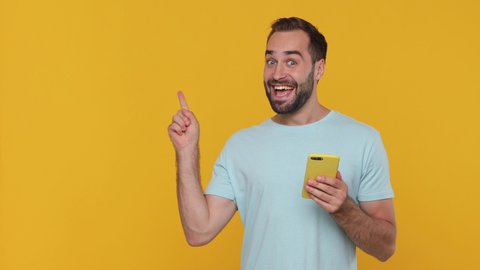 Shocked surprised young guy in basic casual blue t-shirt say wow hold in hand using mobile cell phone pointing fingers aside on workspace isolated on yellow background studio. People lifestyle concept