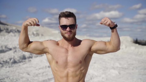 Athletic man posing on camera. Strong bodybuilder without shirt shows his trained body on white canyon background. Muscular sportsman outdoors.