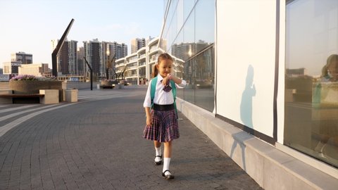 Child girl in uniform is going to school and talking on her pink wrist smart watches and laughing in urban landscape, front view, steadicam shot.