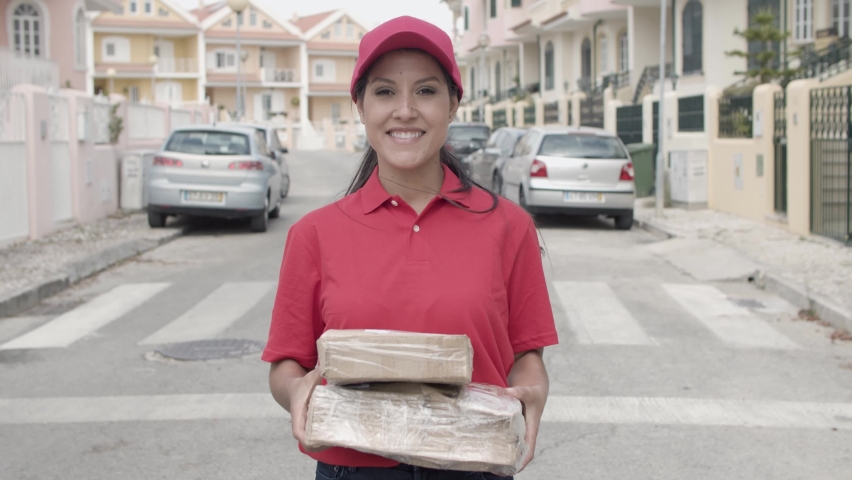 Happy courier girl in red uniform holding packages, standing and posing outside, looking at camera and smiling. Medium shot, front view. Shipping or delivery service concept