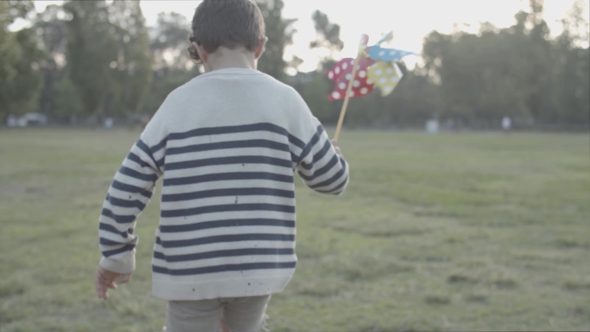 Back view of little boy running on lawn with paper fan. Playful Caucasian child playing in city park and holding colorful swirling toy on stick. Childhood, weekend and summer vacation concept