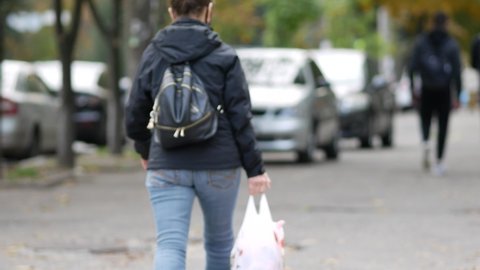 Unrecognizable passers-by are walking down the street. Defocused image. A middle-aged woman with a white plastic bag carries groceries. Backpack. Back view. Autumn day