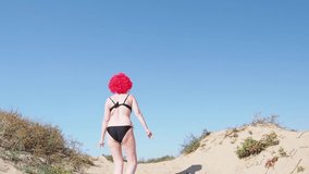 Extravagant flirtatious woman wearing red wig, black shiny swimsuit going over the hill while walking at the beach over sky background - video in slow motion