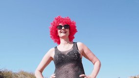 An extravagant flirtatious woman wearing a red wig, a black mesh tunic will remove the devil's horns from his head while walking at the beach over sky background - video in slow motion