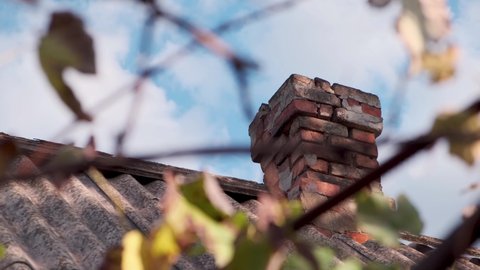 Old vintage brick chimney on a tiled roof on a cloudy sky background at sunny day. Bottom view through foliage.