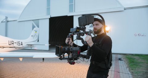 Handsome professional cameraman shooting movie scene on set outdoor. Caucasian bearded man recording video on camera near small airpane. Woman making video on cellphone. Filmmaking industry. Backstage