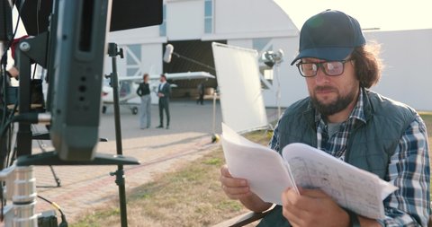 Portrait of filmmaker reading script outdoors on set. Handsome Caucasian male movie director in glasses and cap looking through papers on backstage. Movie production. Filming concept
