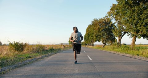 African American handsome strong male jogger in headphones running on road in countryside on summer day. Attractive sporty man jogging outdoor. Sport concept. Sportsman runner. Morning nature.