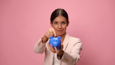 smiling young woman in pink suit putting coin in piggy bank, nodding, making thumbs up gesture on pink background