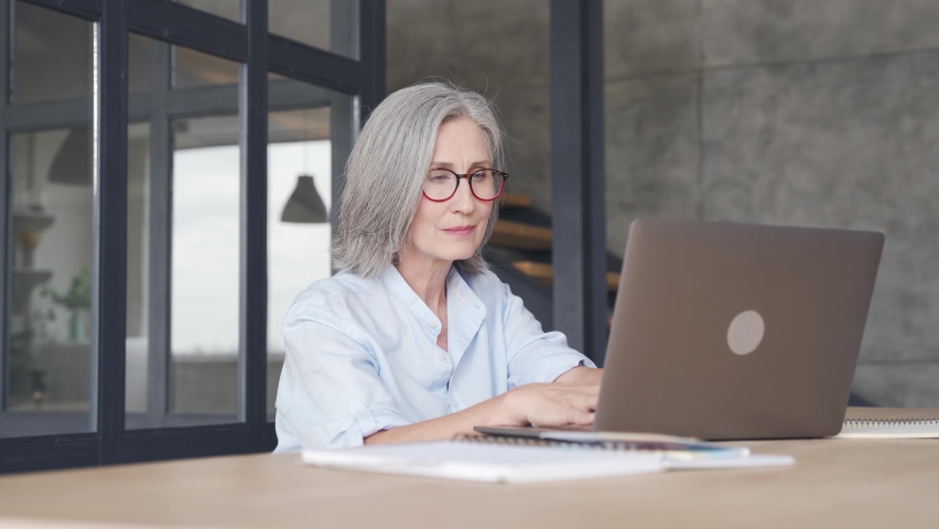 Smiling professional mature business woman wears glasses using laptop computer sits at workplace desk. Happy senior older employee 60s businesswoman executive working typing on pc at home from office. Royalty-Free Stock Footage #1061232397
