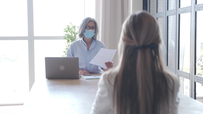 Old hr manager checking female job applicant cv resume at job interview meeting. Senior employer wearing face mask reading seeker employment application. Social distance and safety at work concept. Royalty-Free Stock Footage #1061232403