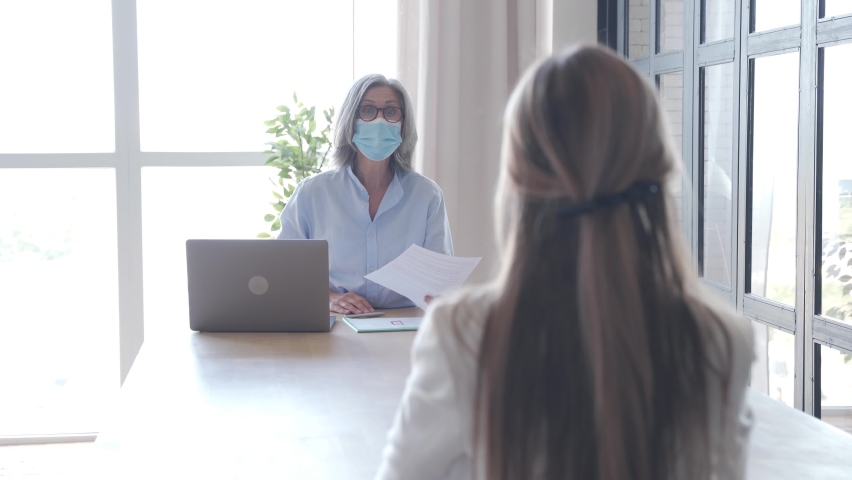 Old hr manager checking female job applicant cv resume at job interview meeting. Senior employer wearing face mask reading seeker employment application. Social distance and safety at work concept. | Shutterstock HD Video #1061232403