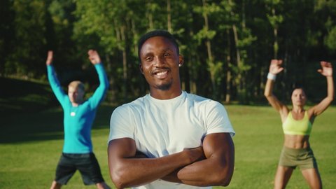 Confident afro-american bodybuilder with crossed arms smiling cheerfully at camera on the sportsground. Personal trainer portrait. Active people are exercising.