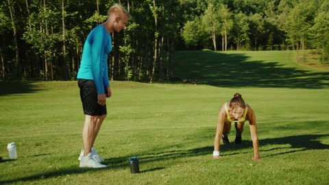 Active pretty sportswoman doing highly intensive physical exercises outdoors. With help of personal trainer athletic girl performing squats and jumps on the grass. Fitness couple.