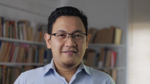 Portrait of cheerful Asian male librarian, looking at camera and smiling, man standing against books in library