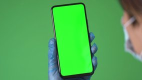 Female hand in medical gloves holding the smartphone on green screen chroma key background. Mobile phone mock-up for your product.