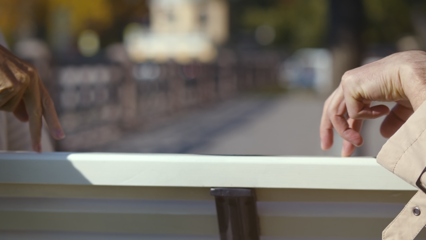 Close up of aged couple moving fingers on wooden bench back outdoors. Romantic mature man and woman holding hands relaxing on bench over blurred city background