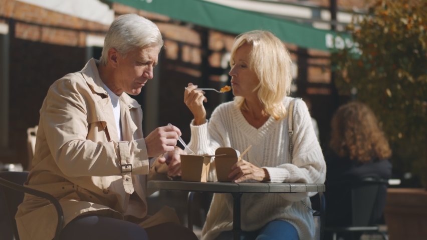 Middle-aged couple sitting at small outdoor cafe table and enjoying takeaway lunch. Portrait of senior husband and wife relaxing on restaurant terrace eating chinese wok on warm autumn day Royalty-Free Stock Footage #1061239795