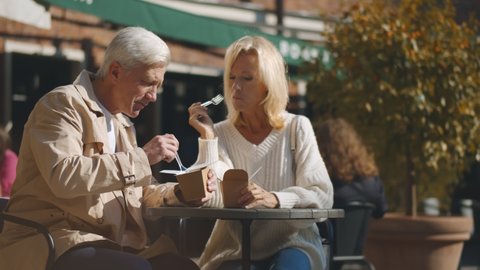 Middle-aged couple sitting at small outdoor cafe table and enjoying takeaway lunch. Portrait of senior husband and wife relaxing on restaurant terrace eating chinese wok on warm autumn day