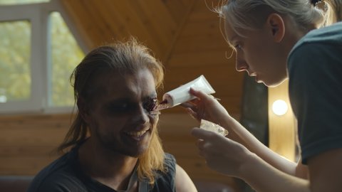 Young woman make-up artist using special equipment applying complex zombie make up on male face. Man model during process of creating zombie face