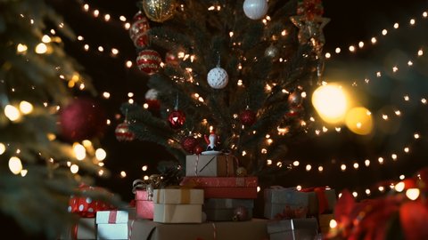 Santa claus figurine with stack of gift boxes under a beautiful decorated christmas tree against bokeh lights at night.