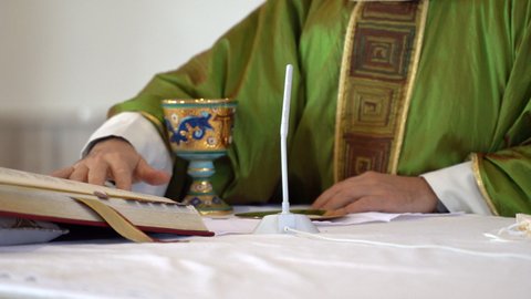 Priest in green cassock standing next to church altar and reading Holy Scripture turning pages. Local pastor performing sermon on celebration mass and praying to God with people. Religion and catholic