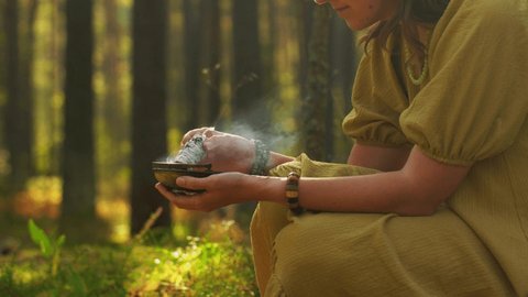occult science and supernatural concept - young woman or witch with smoking white sage performing magic ritual in forest