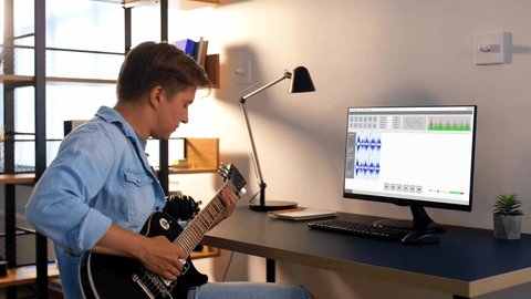 leisure, music and technology concept - young man or musician playing guitar and recording sound with computer program at home studio