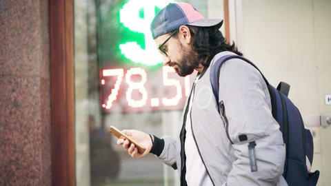 Young handsome man using his smartphone on the street against the background of a led scoreboard with the exchange rate.