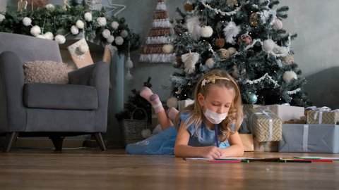 Little Girl in Protective Mask Drawing with Pencils on a Floor near Christmas Tree. New Normal, Virus and Illness Protection, Home Quarantine. Christmas in Covid-19 Pandemic Concept