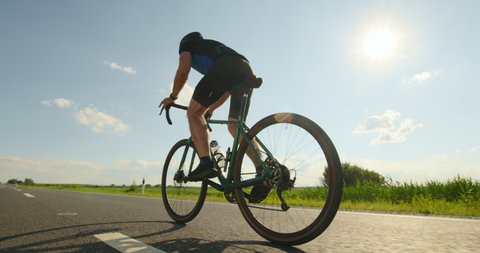 A sportsman on a bicycle is standing on the pedals and developing great speed. Training on a bicycle. Shooting from behind. Sun in the background. 4K