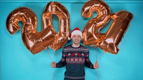 Happy young adult man in santa hat holding golden balloons with numbers on blue
