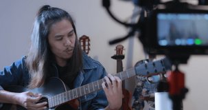 Asian male vlogger performing music and singing show to streaming digital internet online audience listening at home. Man who have long hair plays guitar and sing a song in slow motion shot.