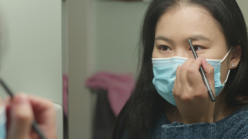 Young Asian American woman with face mask applying eyebrow makeup